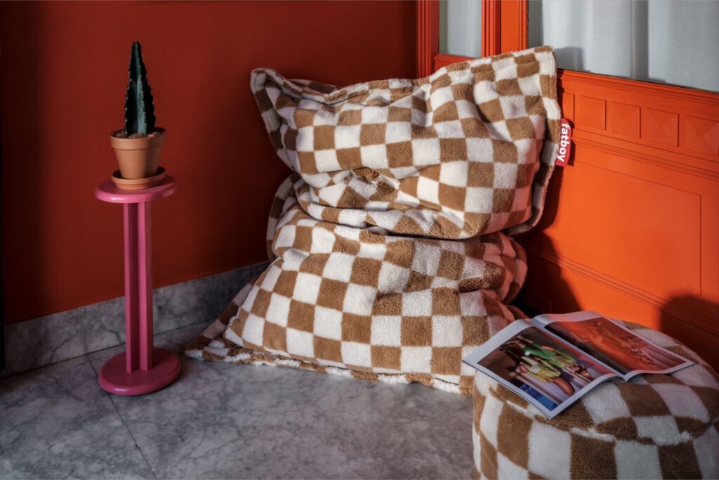 Un pouf Fatboy Point Teddy Chess Almond Creme est posé sur un sol en marbre à côté d'un ottoman rond, d'une table d'appoint rose avec un cactus en pot et d'un magazine ouvert dans une pièce aux murs orange.