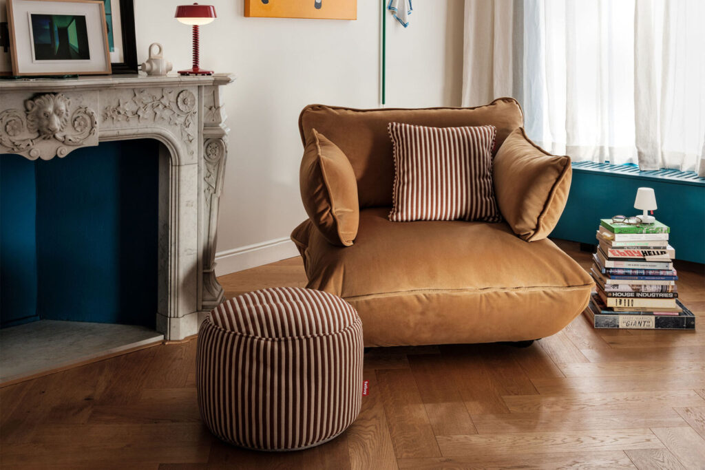 Un grand fauteuil beige avec des coussins rayés est posé sur un sol en bois près d'une cheminée en marbre ; un ottoman Fatboy Point Terry Cognac Creme, une pile de livres et une fenêtre sont également visibles.