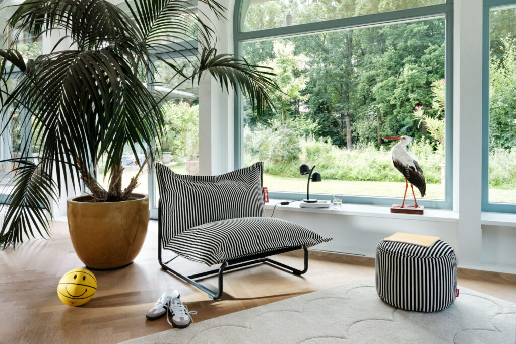 Un espace de vie moderne avec une chaise longue rayée, un ottoman Fatboy Point en Terry Cognac Creme, un grand palmier en pot, une balle au visage souriant, des chaussures et une cigogne taxidermique près de grandes fenêtres donnant sur de la verdure.