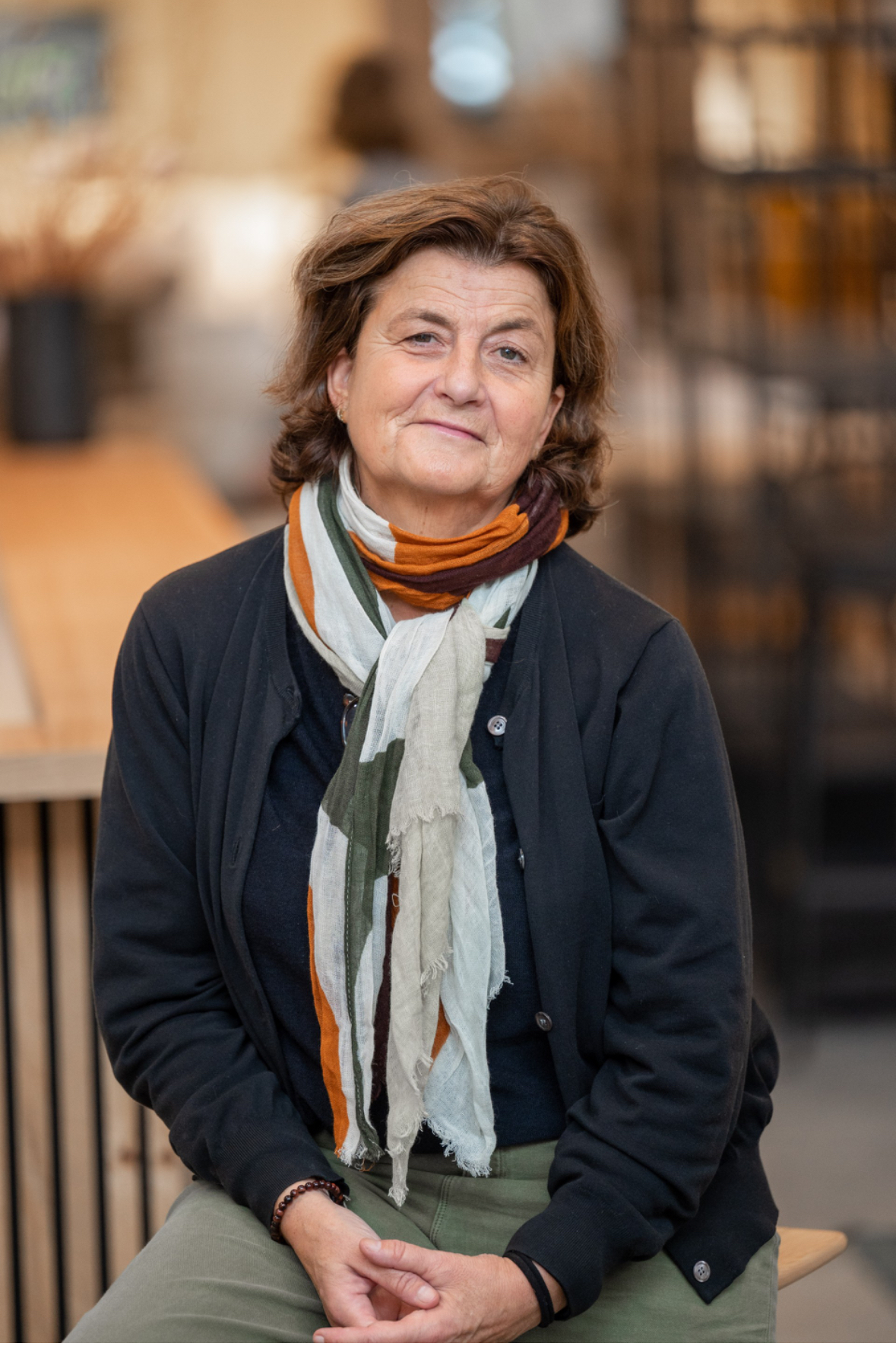 Une femme d'âge moyen aux cheveux bruns est assise à l'intérieur, vêtue d'un pull sombre, d'un pantalon vert et d'une écharpe multicolore - dégageant la confiance calme que l'on voit souvent chez les professionnels - tandis qu'elle regarde calmement la caméra.