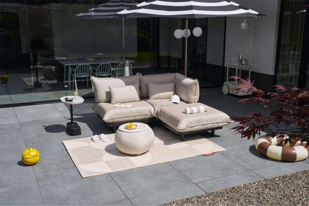 Un patio extérieur moderne présente l'ensemble Paletti Small sectionnel en Royal Brulee, un ottoman rond, des petites tables, un parasol noir et blanc, un ballon de basket et des flotteurs de piscine sur une surface carrelée.