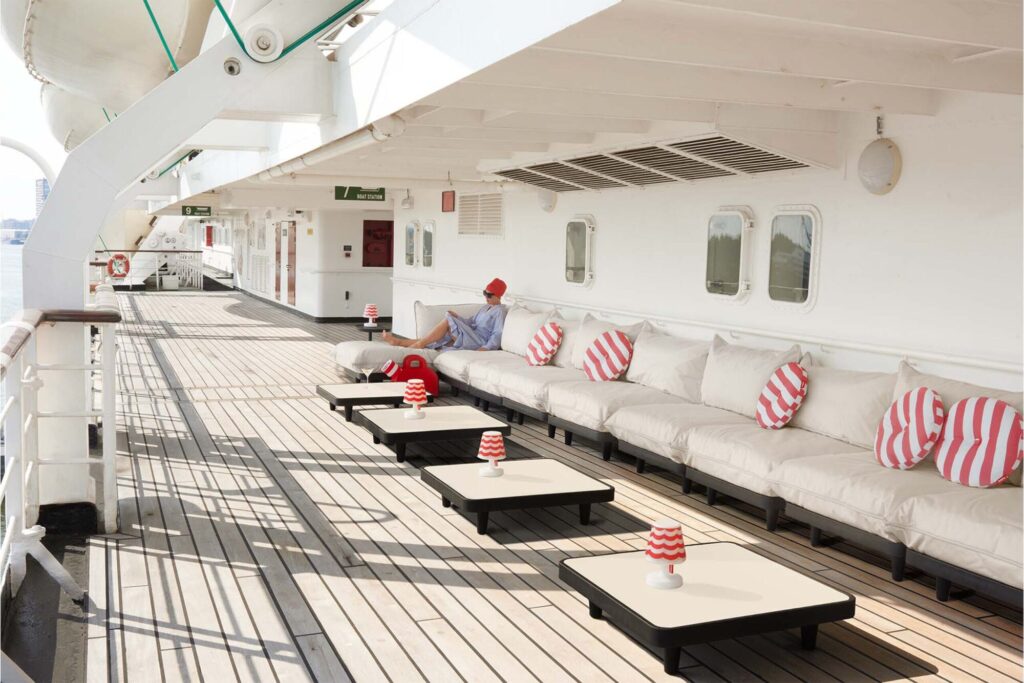 Une personne coiffée d'un chapeau rouge se détend sur une longue banquette blanche Paletti Set Medium avec des coussins rayés sur le pont d'un bateau de croisière, à côté de tables basses avec des lampes rayées.