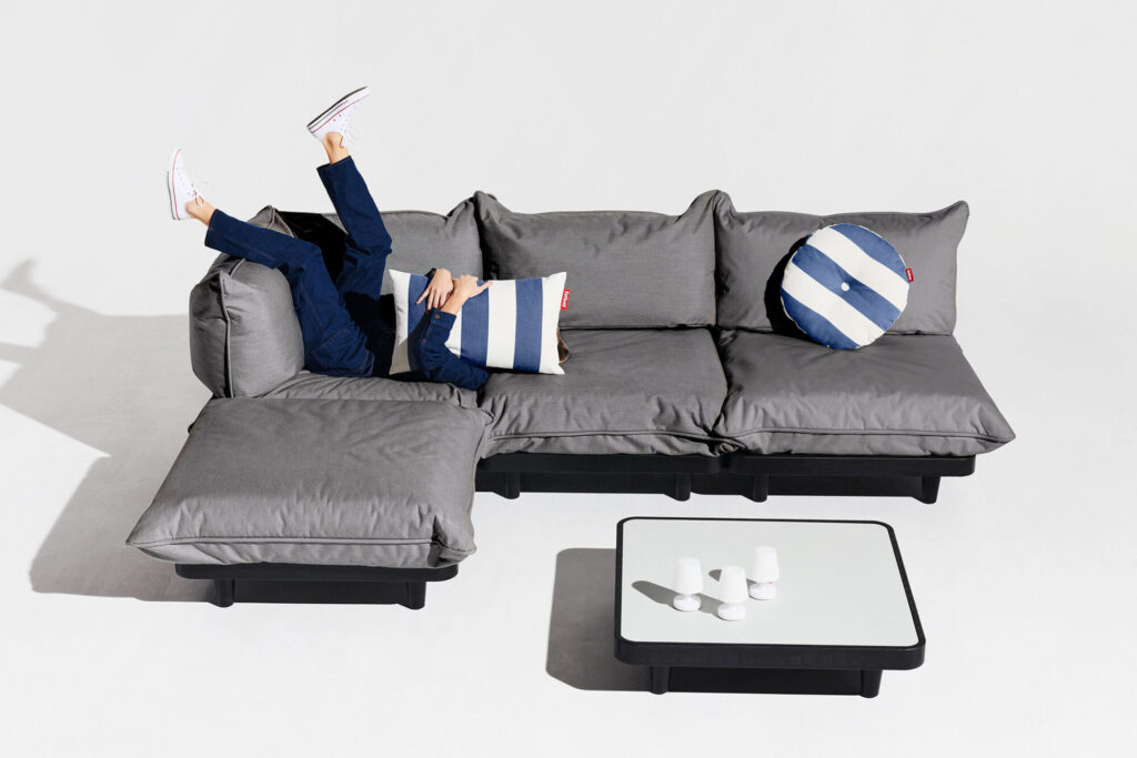 Une personne se prélasse sur un canapé sectionnel moderne de couleur gris roche, les jambes relevées, se couvrant partiellement le visage d'un oreiller rayé ; devant elle se trouve une petite table basse - qui fait partie de l'élégant ensemble Paletti - garnie de tasses.