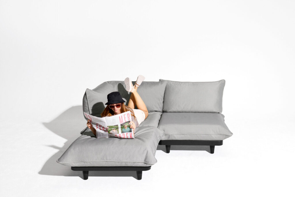 Une personne portant un chapeau et des lunettes de soleil se prélasse sur un canapé Royal Wolf Grey Paletti Set Medium, lisant un magazine sur un fond blanc uni.