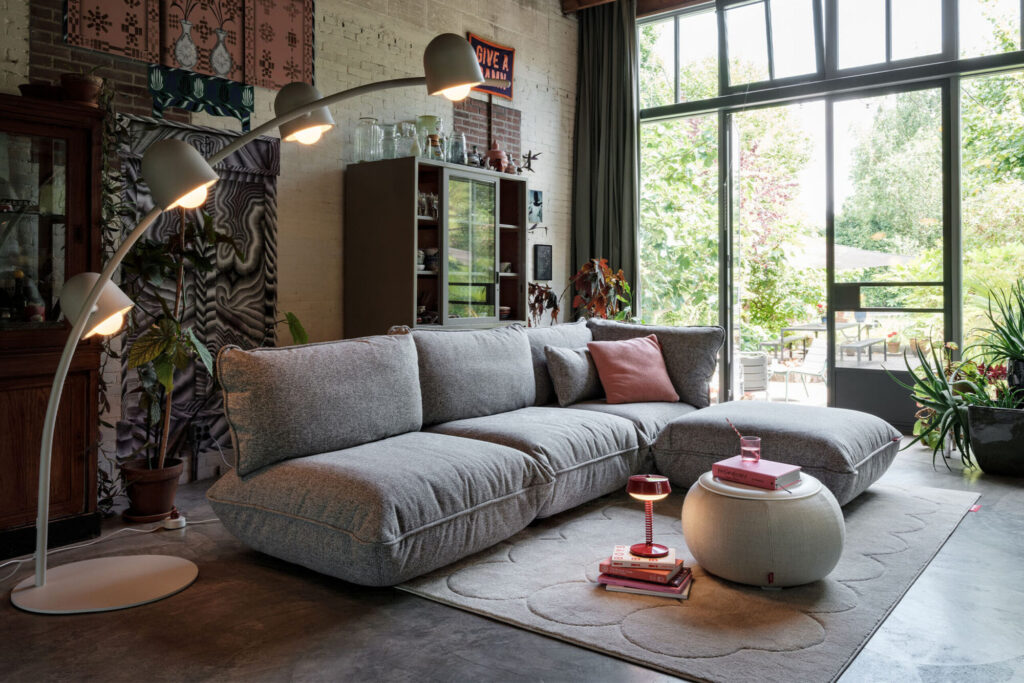 Salon moderne avec un canapé sectionnel Sumo gris, un lampadaire, un ottoman rond et de grandes fenêtres donnant sur un jardin. Des plantes d'intérieur et des œuvres d'art décorent l'espace, tandis qu'une table en marbre Mingle ajoute une touche de sophistication.