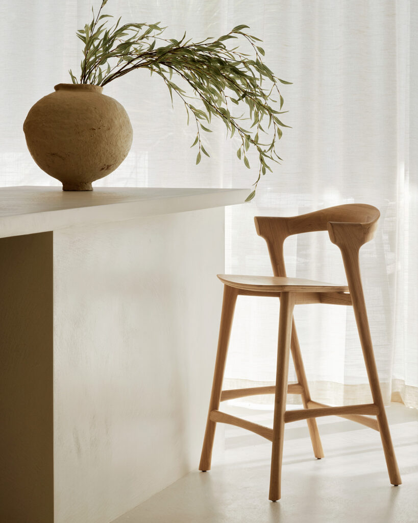Une chaise en bois clair Bok est posée à côté d'un comptoir blanc avec un vase rond beige contenant des branches feuillues, le tout entouré de rideaux blancs transparents.