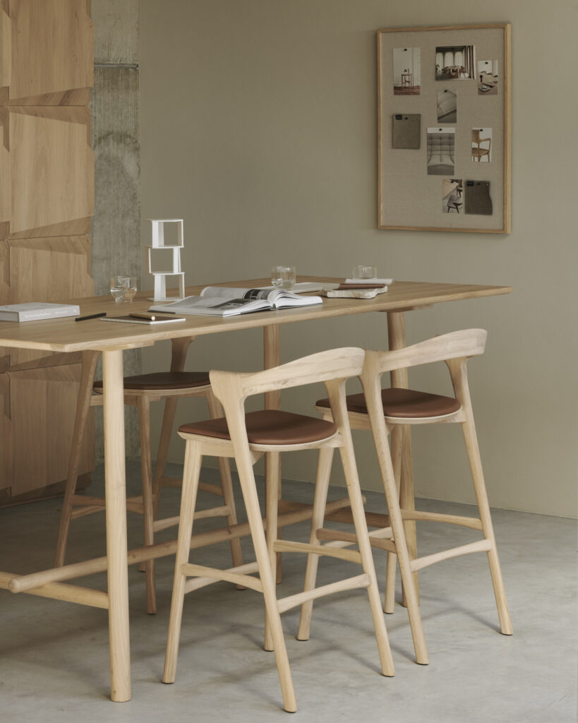 Le coin repas minimaliste est composé de deux chaises Bok en bois clair, d'une table assortie, de livres ouverts et de verres posés sur la surface, et d'un tableau d'affichage avec des photos qui orne le mur beige.