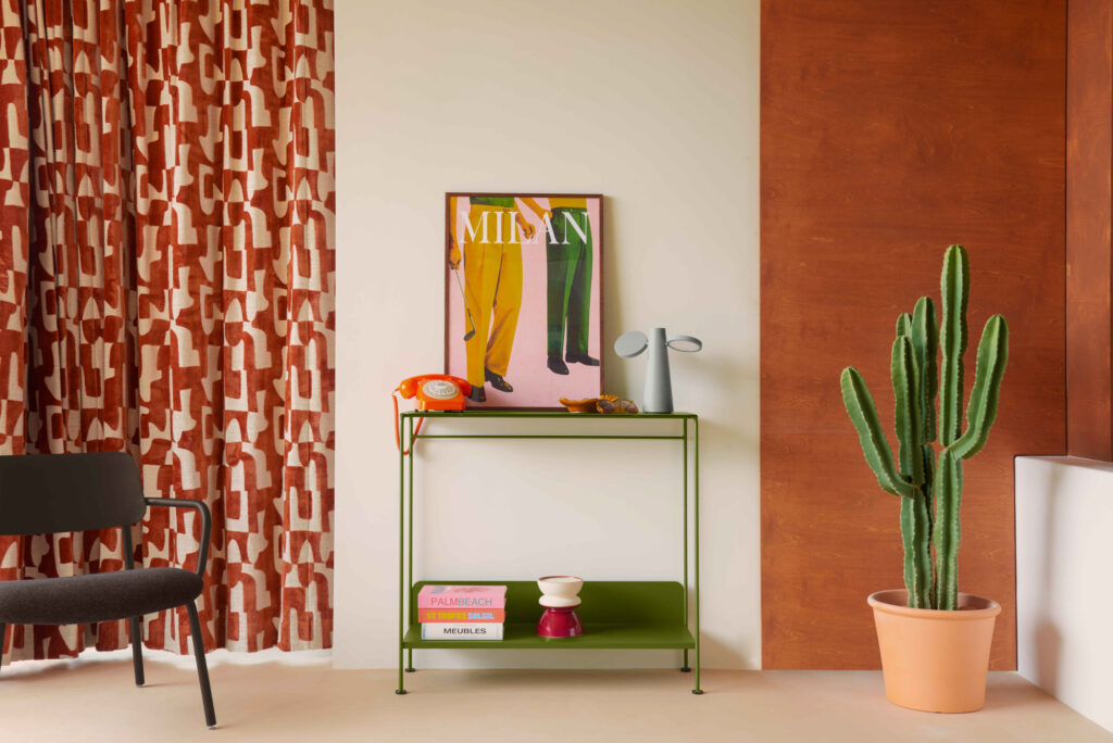 Une petite table verte Console Picolino INDOOR avec des livres, une cafetière rouge et blanche, un vase gris et un téléphone à cadran orange se trouvent contre le mur ; un cactus en pot et des rideaux à motifs évoquent une ambiance Beige Latte confortable.