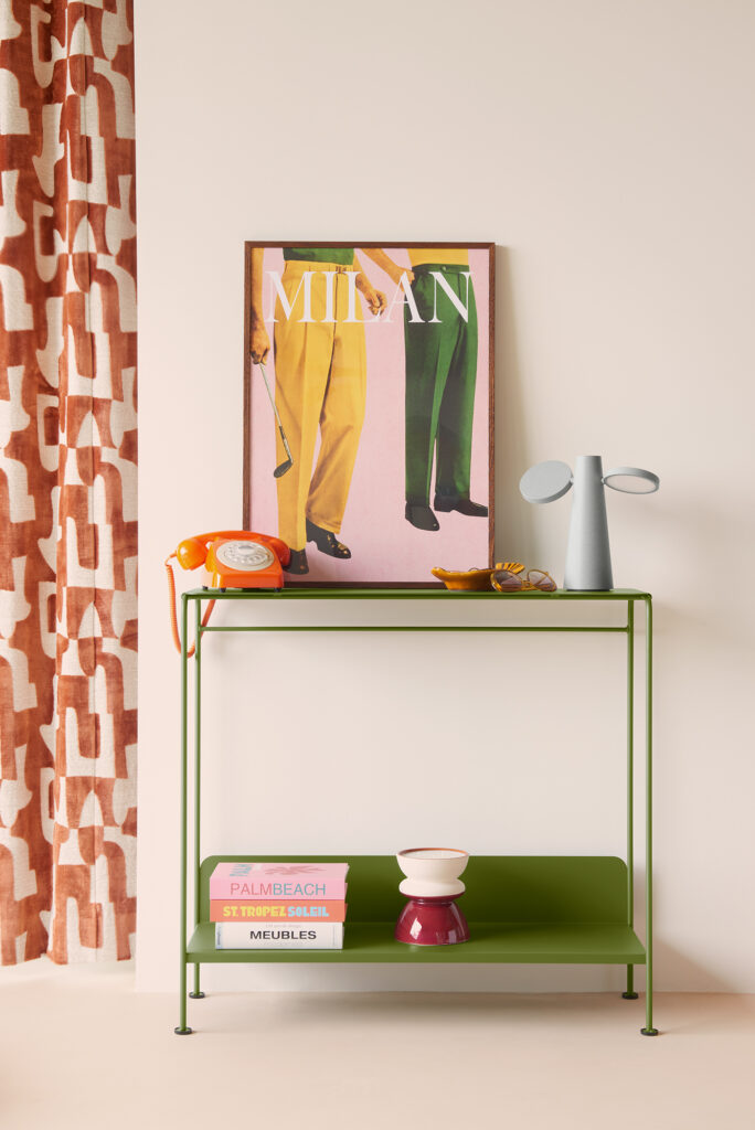 La console Picolino INDOOR en métal vert présente des livres, un bol rouge et blanc, un vase gris, un téléphone à cadran orange et des œuvres d'art encadrées à côté de rideaux à motifs sur un mur blanc.
