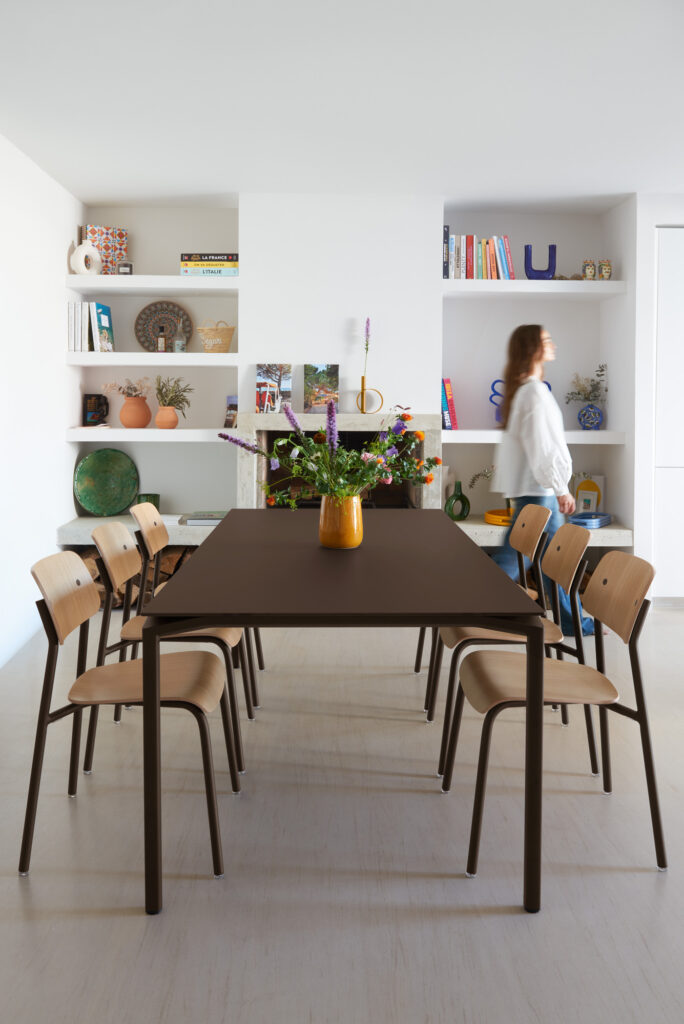 Une salle à manger moderne avec une table rectangulaire foncée, six chaises en bois Chaise Studie, un vase de fleurs, des étagères encastrées avec des livres et de la décoration, et une personne marchant à l'arrière-plan.