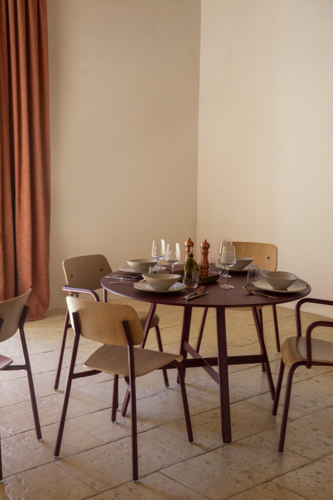 L'ensemble de salle à manger Chaise Studie comprend une table ronde pour quatre personnes avec des assiettes, des verres et des condiments, entourée de chaises en bois de style chaise sur un sol carrelé à côté d'un mur beige et d'un rideau couleur rouille - idéal pour un dîner confortable ou un bureau décontracté.