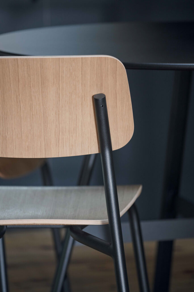 Gros plan sur la Chaise Studie, une chaise en bois avec des pieds en métal noir, placée à côté d'une table sombre dans un décor minimaliste.