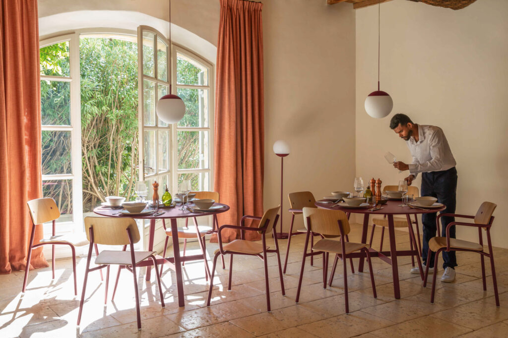 Un homme dresse une table dans une pièce ensoleillée avec de grandes fenêtres, des tables rondes, des chaises en bois et des rideaux orange, créant ainsi un espace accueillant qui rappelle la Chaise Studie.