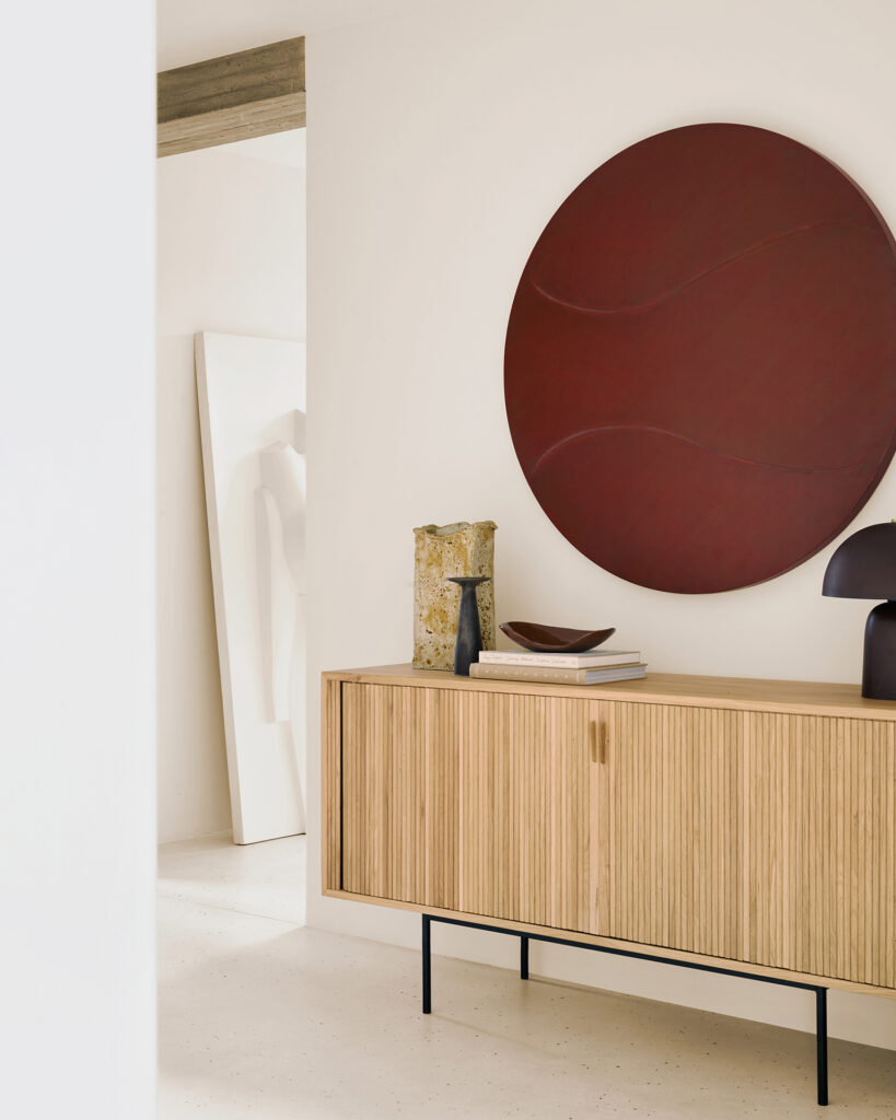 Un buffet moderne en bois Roller-Max en chêne naturel huilé avec des livres, un vase décoratif et une lampe sombre se trouve au-dessous d'un grand art mural circulaire rouge dans une pièce minimaliste aux murs neutres. Longueur : buffet 168 cm.