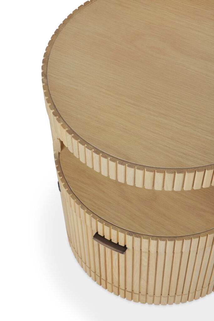 Table basse ronde en bois chêne naturel avec des côtés rainurés, une étagère ouverte et deux poignées rectangulaires découpées sur les côtés opposés. Finition huilée pour une meilleure durabilité, inspirée des éléments de design du Caisson Boomerang.