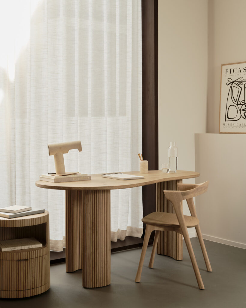 Un espace de travail minimaliste avec un Caisson Boomerang, un bureau en bois chêne naturel huilé, une chaise, une table d'appoint, un carnet de notes et un stylo, une bouteille d'eau et une gravure de Picasso sur le mur, le tout entouré de rideaux transparents.