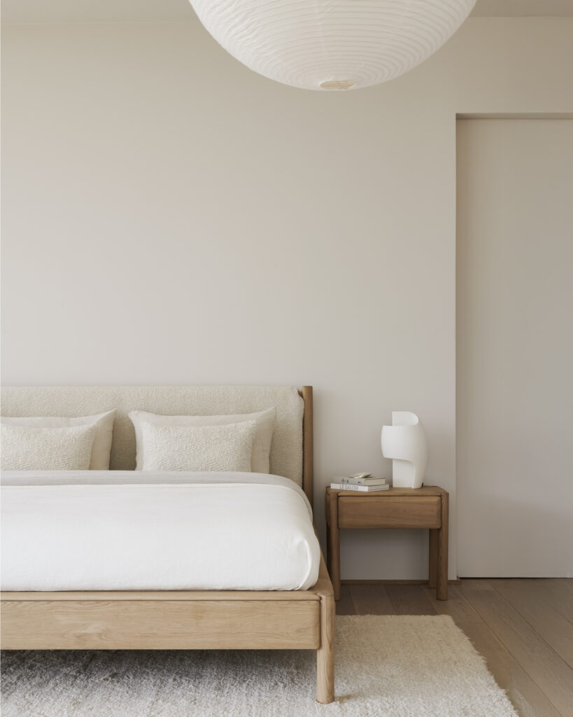 Chambre à coucher minimaliste avec un cadre de lit en bois, une literie blanche, deux oreillers et la table de chevet PI - 1 tiroir contenant une lampe et des livres. Un grand plafonnier rond complète l'ensemble.