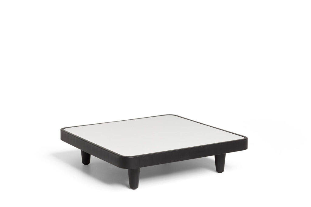 La table Paletti présente un design carré et bas avec un plateau blanc et un cadre noir anthracite, soutenu par quatre pieds noirs courts, sur fond blanc.
