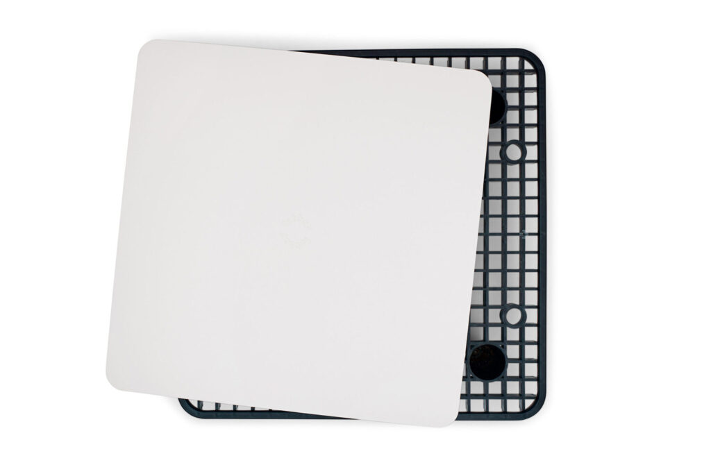 Une planche à découper rectangulaire blanche est placée en diagonale sur le plateau grillagé en plastique noir d'une table Paletti anthracite, vue d'en haut.