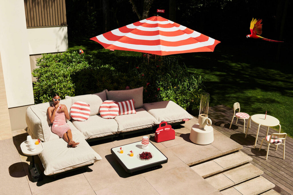 Une personne vêtue de vêtements rayés se détend sur un canapé de terrasse sous un parasol rouge et blanc, avec des boissons, des en-cas et un perroquet volant à proximité. Une table Paletti anthracite avec des chaises se trouve à droite.