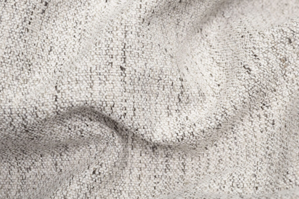 Gros plan sur Mingle Grid Stone, un tissu gris clair texturé avec des fils visibles et des plis doux, idéal pour les tissus d'ameublement Sumo Hocker.