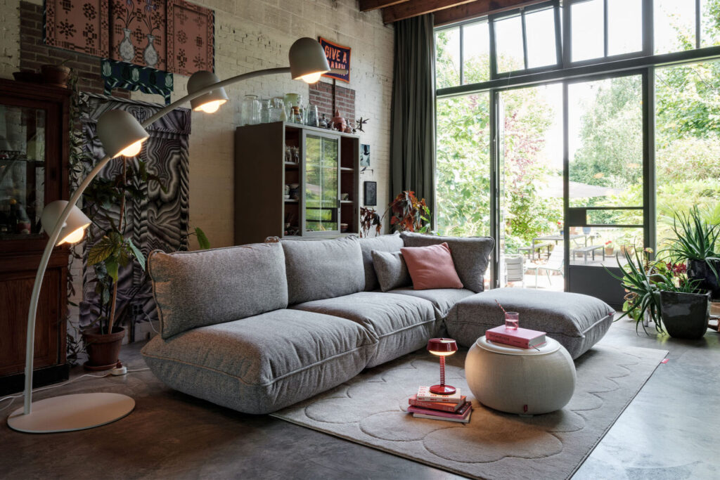 Un salon moderne avec un canapé sectionnel Sumo gris, un ottoman Mingle Marble, un lampadaire, des portes en verre, des plantes d'intérieur et un décor éclectique. La lumière naturelle remplit l'espace.