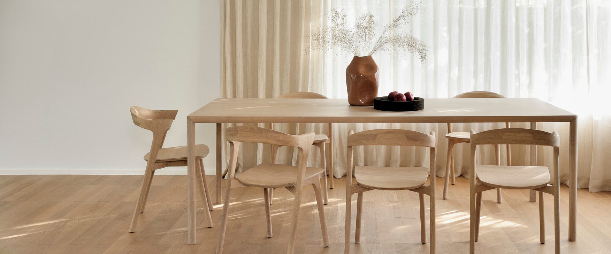 Une salle à manger minimaliste avec une table en bois clair, six chaises assorties et un bol de fruits crée une ambiance sereine 2025 BE-FR, avec des rideaux beiges et une grande fenêtre - parfaite pour votre curseur principal.