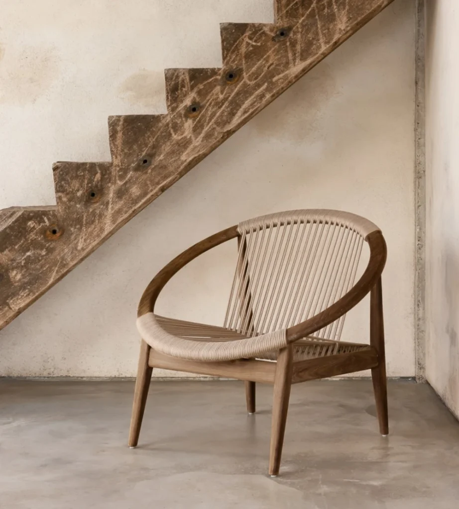 Une chaise moderne en bois avec un cadre circulaire et une assise tissée est posée sur un sol en béton sous un escalier rustique et inachevé - parfait pour votre rafraîchissement du vendredi noir.