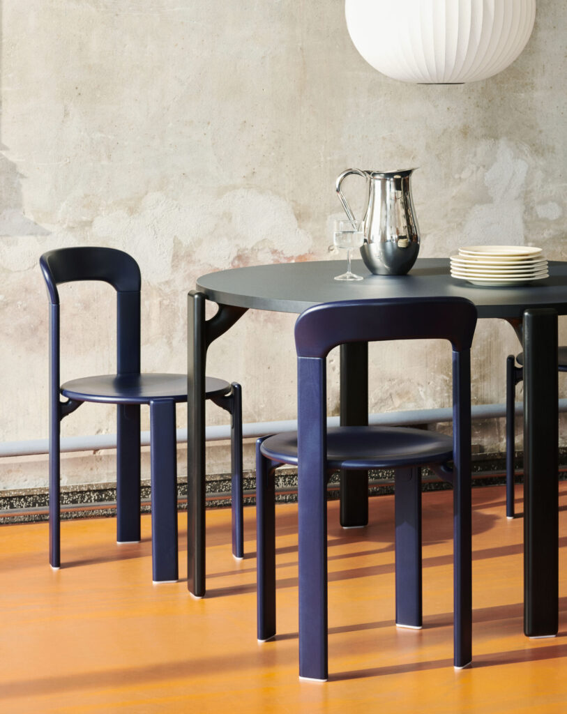 Une table à manger moderne avec deux chaises bleu foncé, un pichet métallique, des assiettes empilées et un verre à vin est placée contre un mur texturé et un sol orange - parfait pour votre mise à jour du Black Friday.