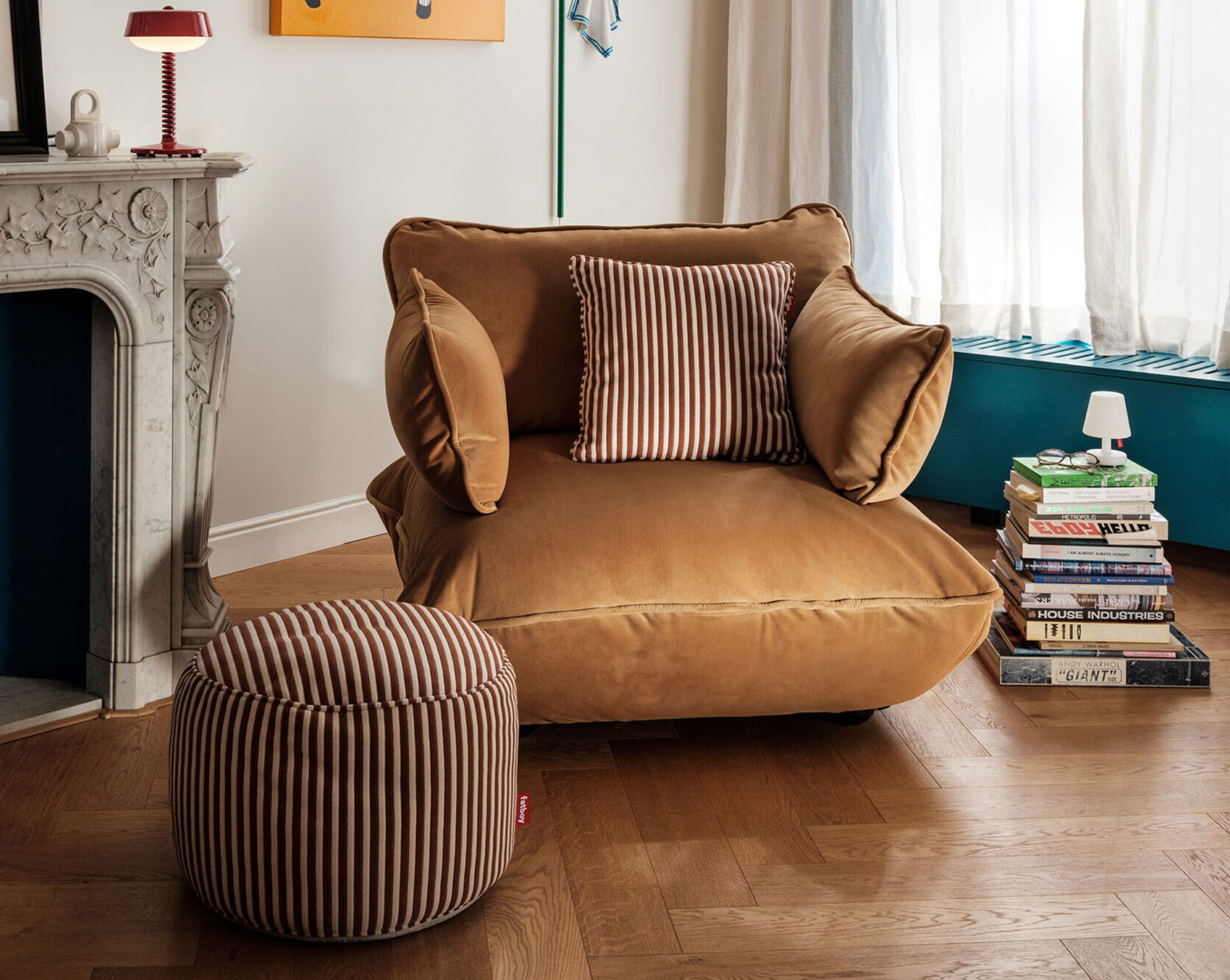 Un fauteuil bronzé en peluche avec des coussins rayés est posé à côté d'un ottoman rayé assorti dans une pièce confortable avec une cheminée en marbre, une pile de livres et un plancher en bois, parfaite pour se détendre après les achats du vendredi noir.