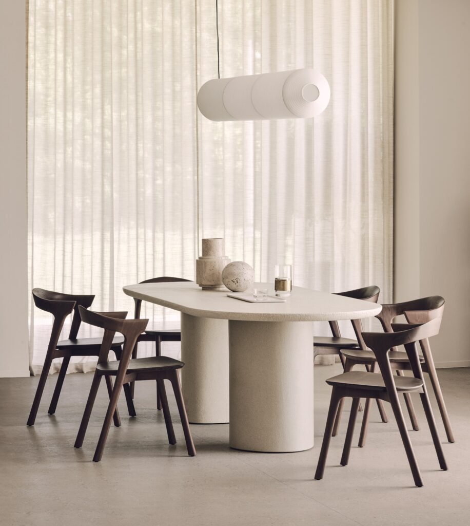 Une salle à manger moderne avec une table ovale beige, six chaises en bois foncé, des objets décoratifs et une lampe suspendue cylindrique blanche, parfaite pour accueillir les réunions du Black Friday, le tout entouré de rideaux diaphanes allant du sol au plafond.
