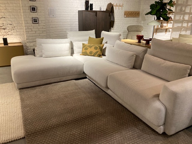 Un salon moderne comprend le canapé sectionnel blanc Olta - Willow (L2Hz+3.5z) C50 3715 Cottage, un tapis texturé beige et des étagères murales, mêlant décor minimaliste et charme cottage sur un mur de briques blanches.