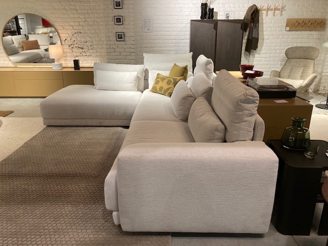 Un salon moderne avec le canapé sectionnel blanc Olta - Willow (L2Hz+3.5z) C50 3715 Cottage, un miroir rond, un buffet en bois, une lampe et des coussins décoratifs contre un mur de briques blanches.