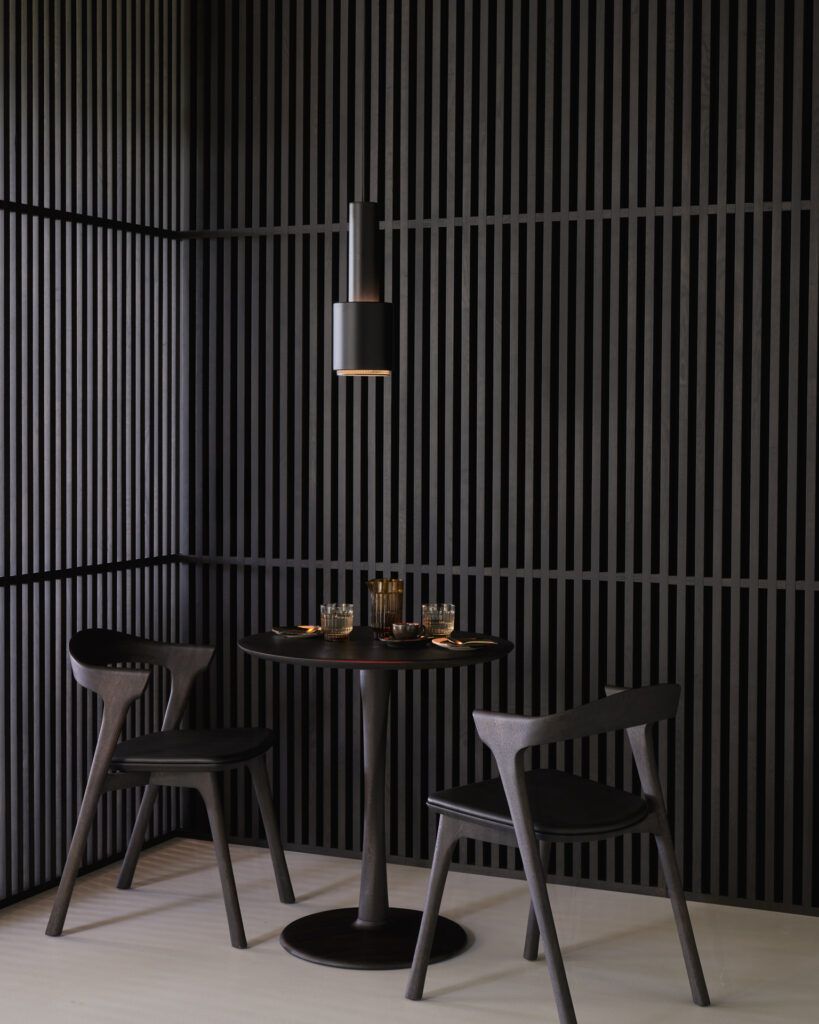 Une table ronde Torsion en chêne teinté avec deux chaises noires est posée dans un coin, avec des verres et des couverts, contre un mur à lattes sombre et une lampe cylindrique suspendue au-dessus.