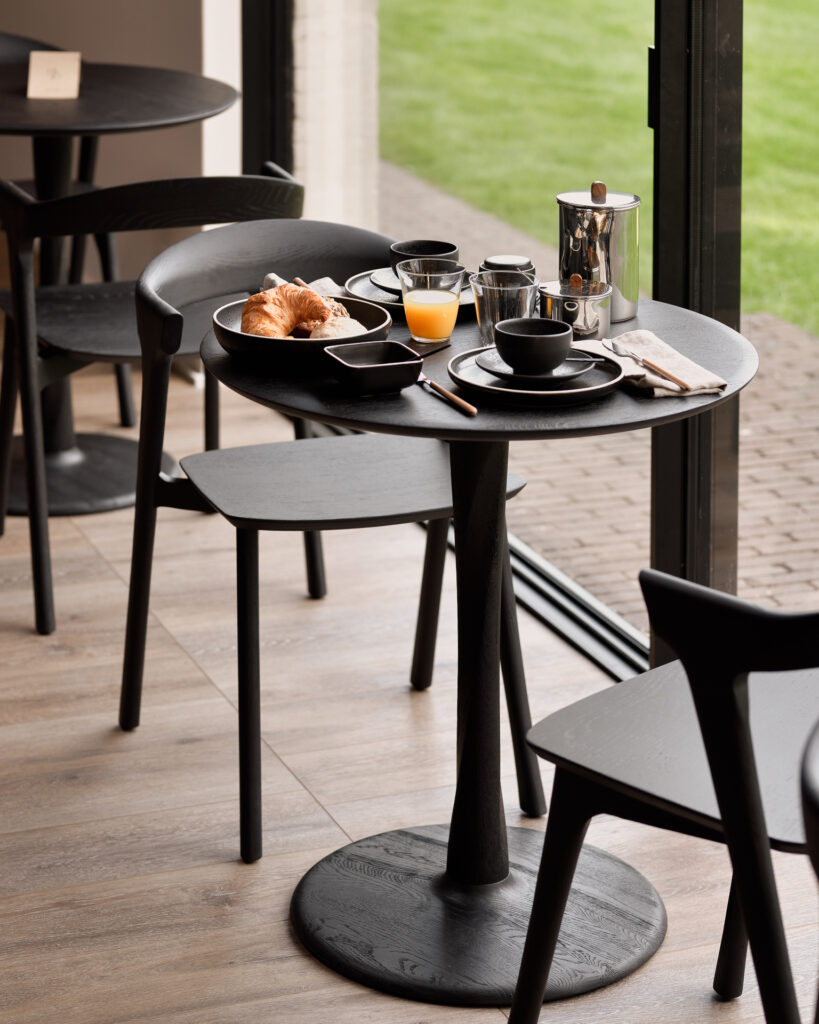 Une petite table ronde Torsion en chêne teinté noir est préparée pour le petit-déjeuner avec des croissants, du jus d'orange, des tasses à café et une presse à café, à côté de portes vitrées ouvertes donnant sur une pelouse.