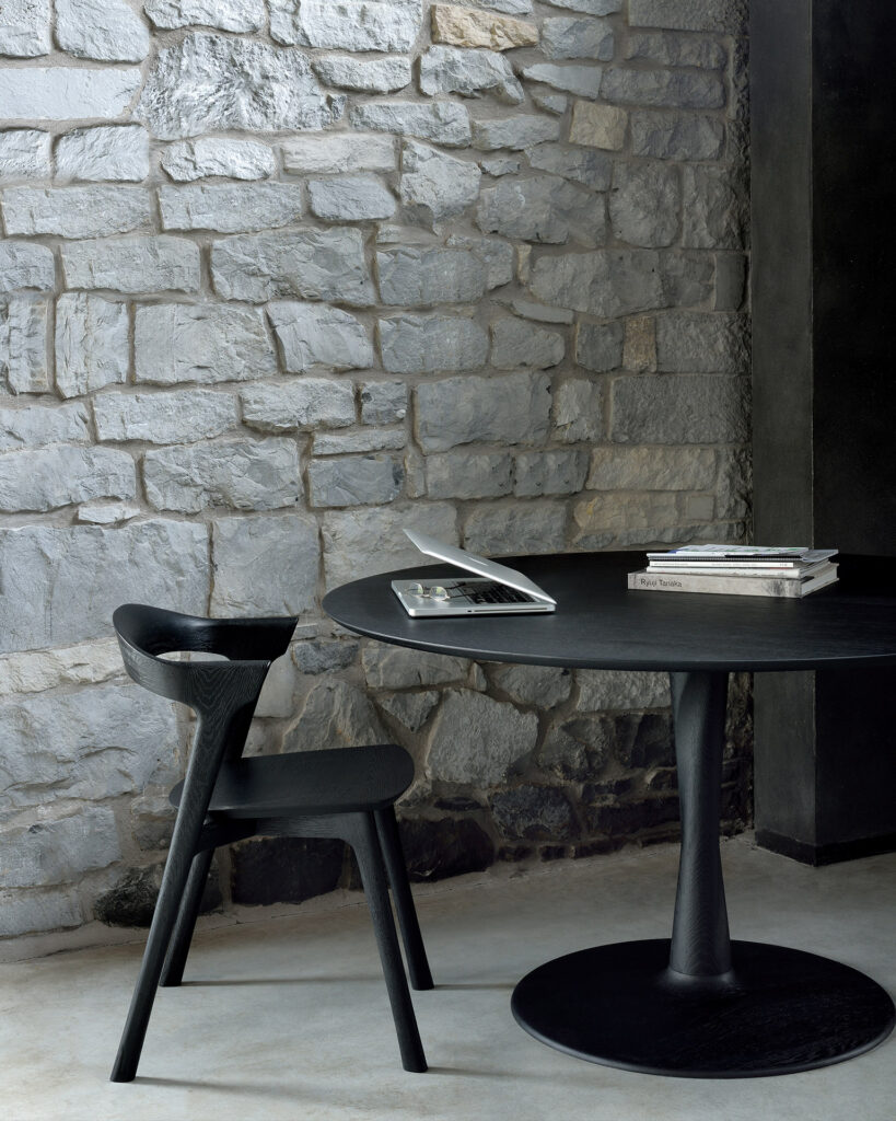 Une chaise noire et une table Torsion en chêne teinté noir avec des livres et un ordinateur portable ouvert sont posés contre un mur en pierre dans un intérieur élégant et minimaliste.