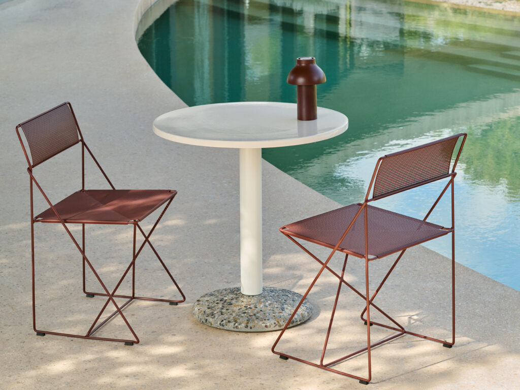 Une petite table ronde blanche avec un vase marron, flanquée de deux chaises X-Line en métal marron, se trouve sur un patio au bord d'une piscine incurvée.