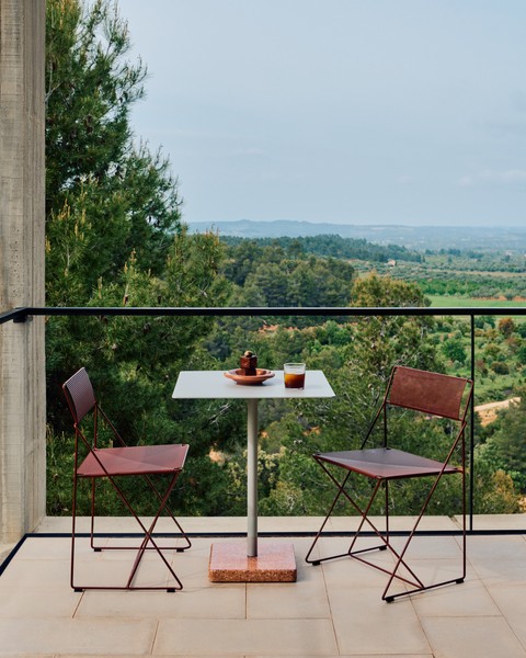 Une petite table de patio avec deux chaises brunes Chaise X-Line surplombe un paysage vert et vallonné ; sur la table se trouvent une assiette, une tasse et un verre.