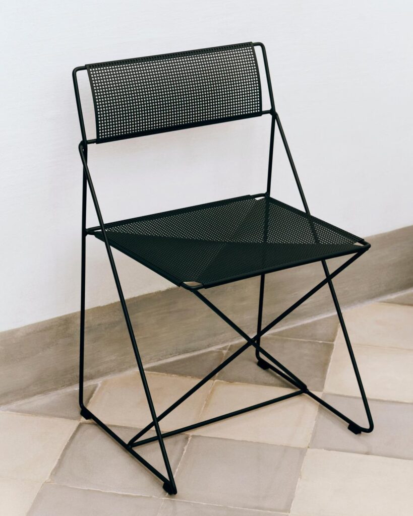 Chaise X-Line chaise pliante en métal noir avec assise et dossier perforés, présentée sur un sol carrelé à côté d'un mur blanc.