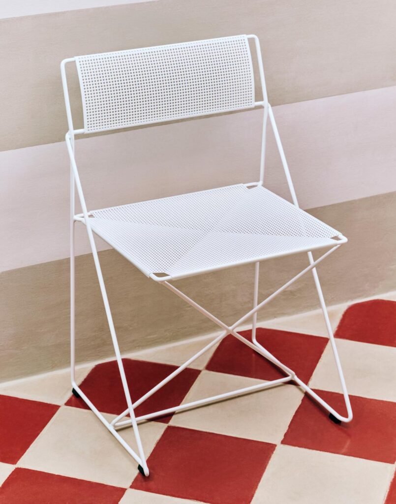 La chaise pliante Chaise X-Line blanche, dotée d'une assise et d'un dossier perforés, est placée sur un sol carrelé à carreaux rouges et blancs, à côté d'un mur à rayures beiges.