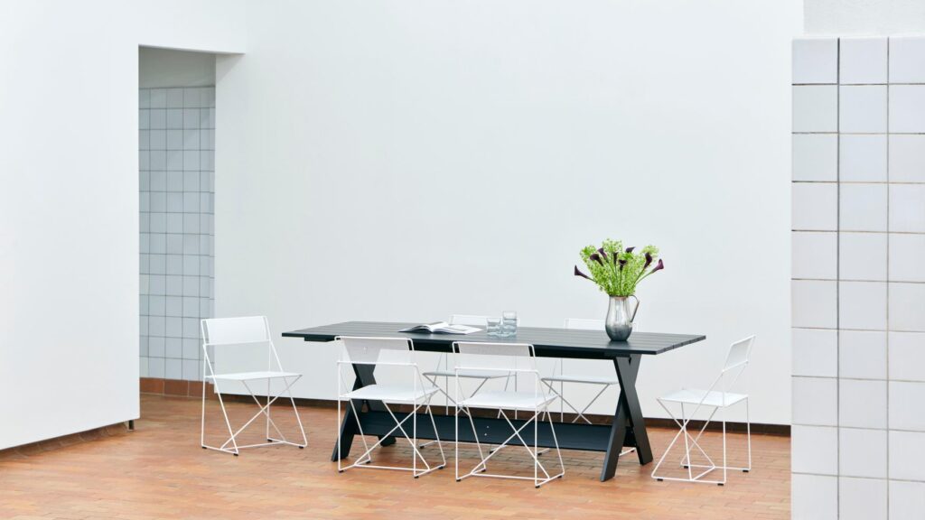 Le coin repas minimaliste se compose d'une table noire, de cinq chaises blanches Chaise X-Line, d'un vase de fleurs et de murs carrelés blancs sur un sol en terre cuite.