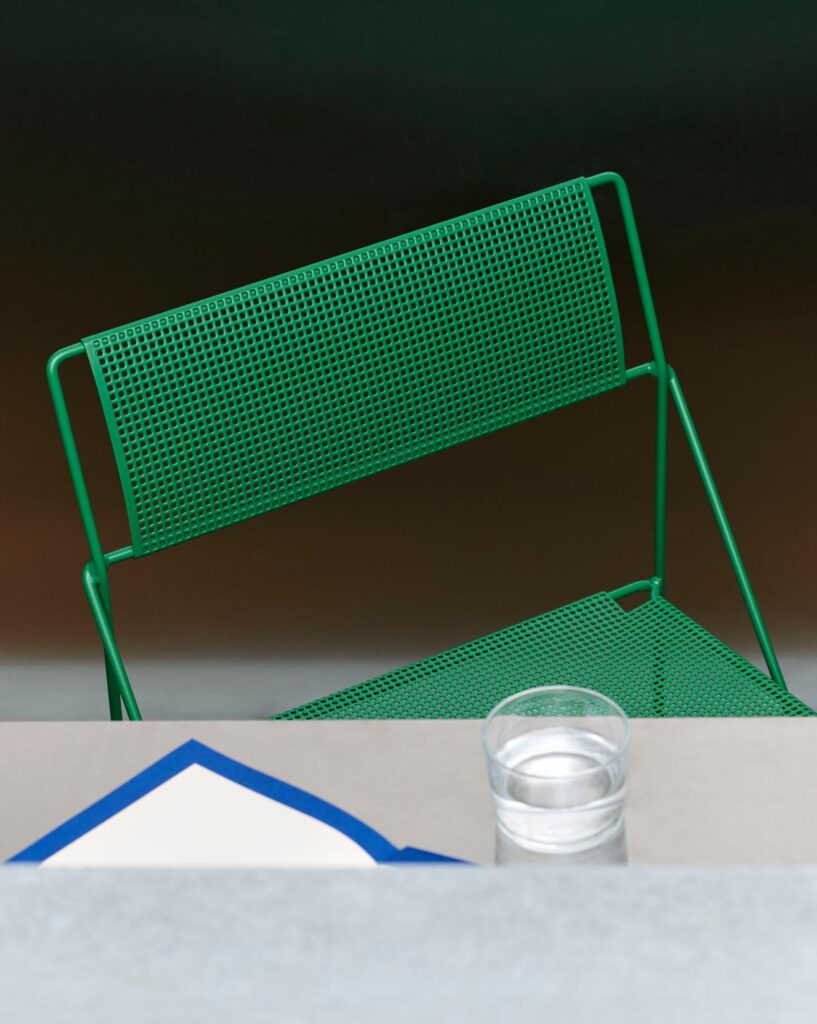Une chaise verte Chaise X-Line en métal perforé est assise derrière une table où se trouvent un verre d'eau et une serviette blanche à bordure bleue.