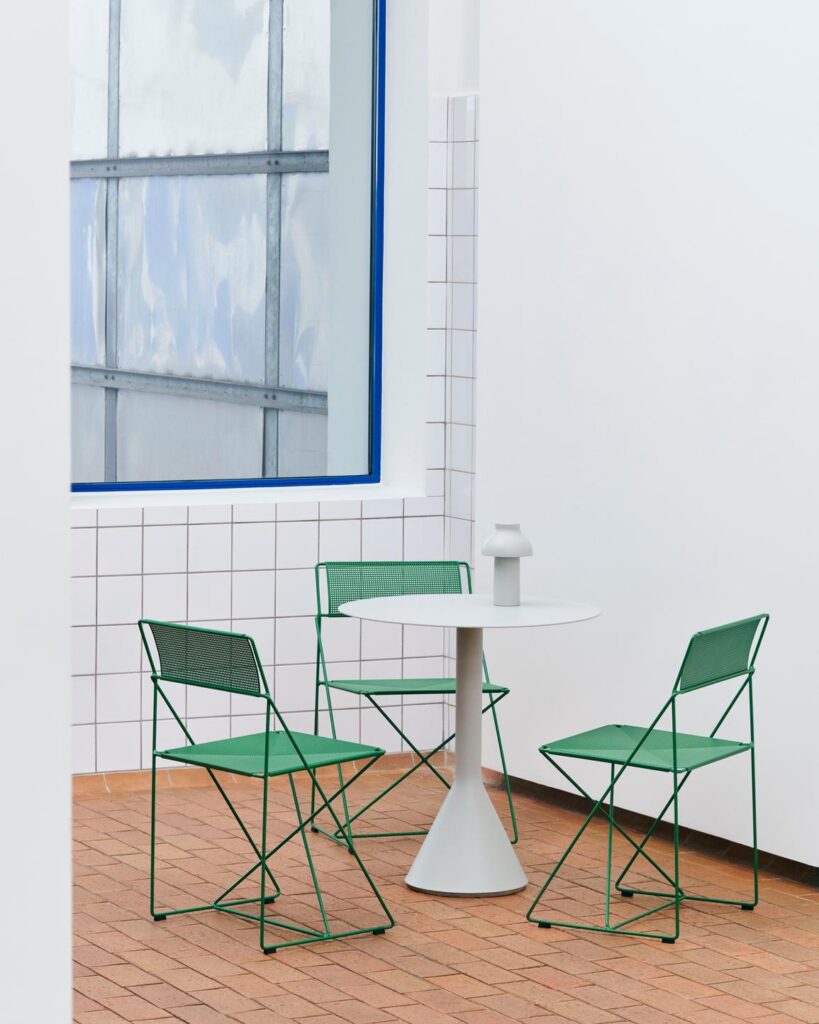 Une table ronde blanche et trois chaises Chaise X-Line vertes en métal sont posées sur un sol carrelé près d'une fenêtre et de murs carrelés blancs dans une pièce minimaliste.