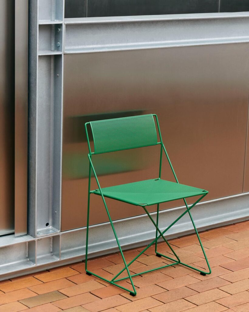 La Chaise X-Line, une chaise pliante en métal vert avec un dossier et une assise en maille, se trouve contre un mur métallique sur un sol en briques.