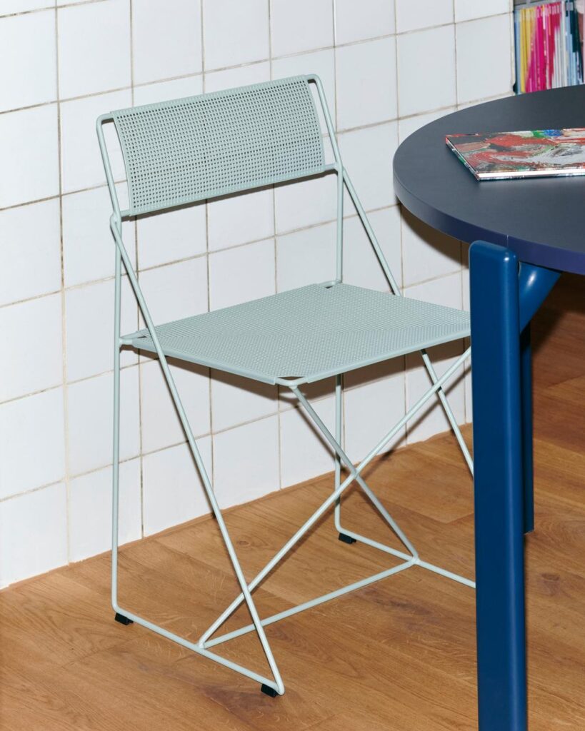 La Chaise X-Line, une chaise pliante en métal gris clair avec une assise et un dossier perforés, est placée à côté d'une table ronde bleue sur un sol en bois devant un mur carrelé.