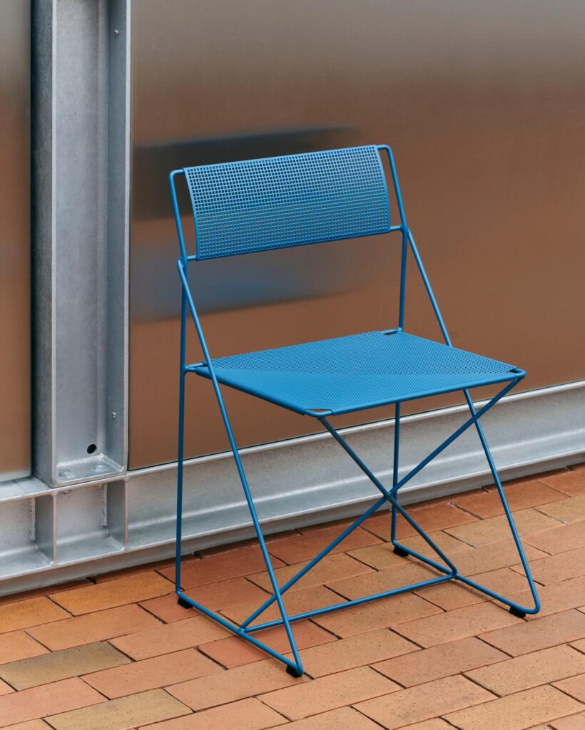 Une chaise pliante Chaise X-Line en métal bleu, dotée d'une assise et d'un dossier perforés, est posée sur un sol en briques brun-rouge, contre un mur métallique.
