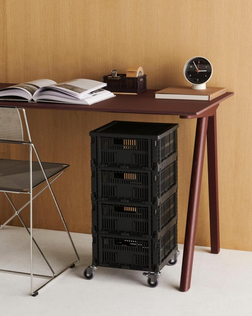 Un bureau surmonté d'un livre ouvert, d'une horloge et d'un distributeur de cassettes ; la chaise X-Line Chair Chrome à côté ; et un chariot de rangement noir à quatre tiroirs sur roulettes en dessous.