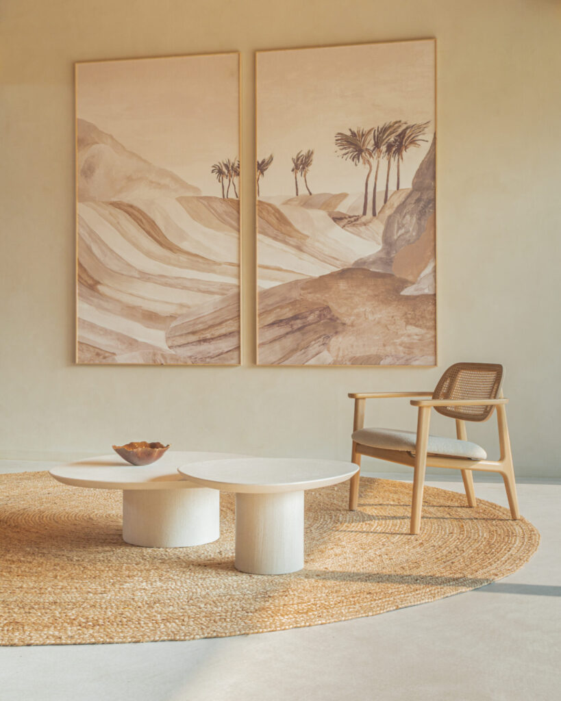 Salon minimaliste avec deux grandes peintures de paysages désertiques, un tapis tissé, une chaise en bois avec coussin, deux tables basses rondes en Ari et un bol décoratif.