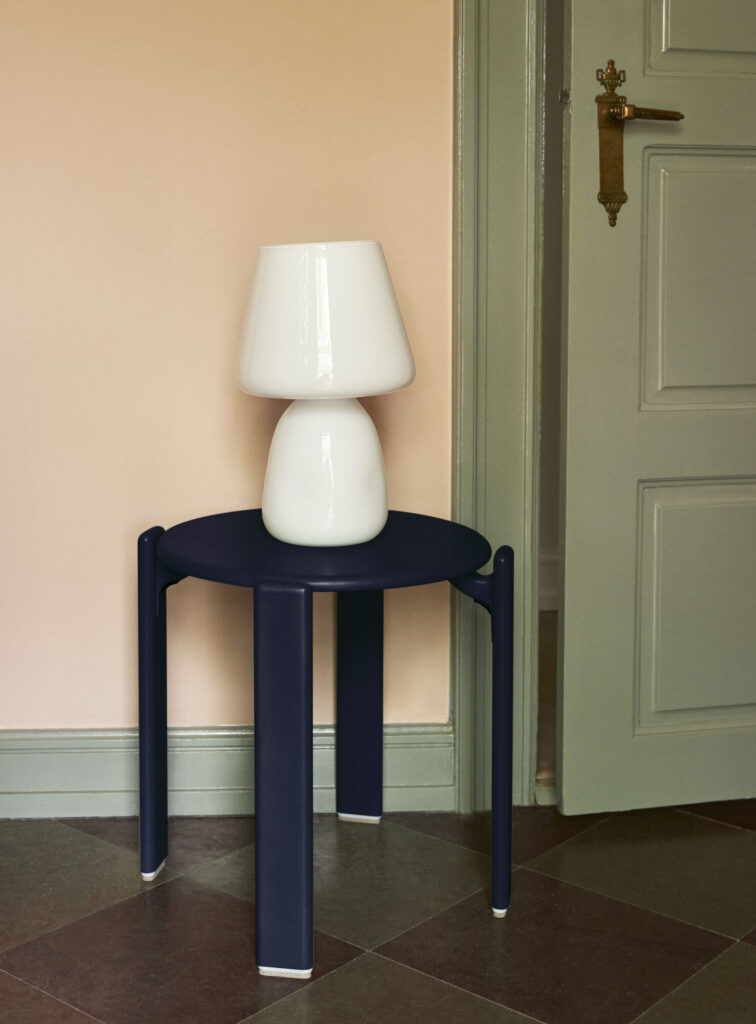 Une lampe de table blanche est posée sur un tabouret Tabouret Rey rouge raisin à côté d'une porte verte dans une pièce aux murs couleur pêche et au sol carrelé.