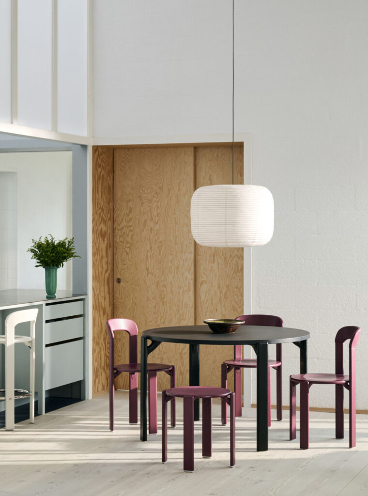 Un coin repas moderne avec une table ronde noire, quatre chaises Grape Red Tabouret Rey, une grande lampe suspendue blanche et une porte coulissante en bois à l'arrière-plan.